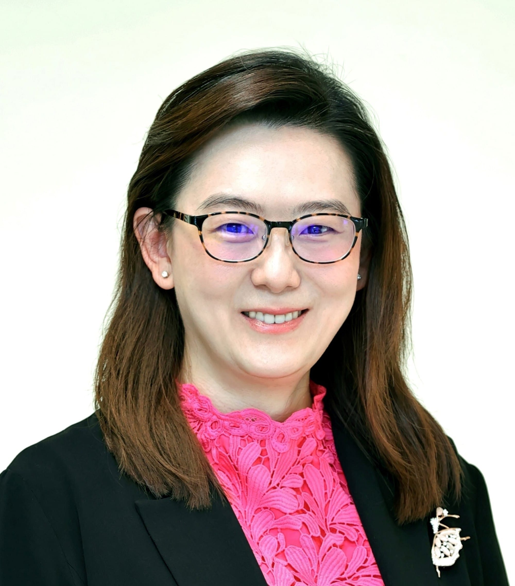 Woman with brown hair and tortoiseshell glasses, wearing a black blazer over a pink lace top, smiling at camera.